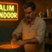 An elderly Indian man cooking at a tandoor oven in a rustic kitchen, under a neon sign that reads "Salim Tandoor".