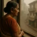 Elderly Indian woman in a saree gazes out of a window onto a quiet, foggy street, reflecting solitude and resilience.