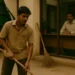 A young South Asian sanitation worker sweeps outside an office while a man in formal attire observes him from inside through a glass window.