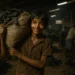 A young boy in Dharavi carries a heavy sack of scrap leather, smiling despite hardship inside a dim leather tanning workshop.