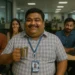 Laughing coworkers surround a cheerful, chubby Indian man with coffee in tech office